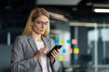 Ofis ortamındaki olgun iş kadını iletişim ve yönetim görevleri için akıllı telefonu kullanıyor. Kendinden emin ve odaklanmış görünüyor. Liderlik ve profesyonellik gösteriyor..