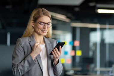 Ofisteki olgun iş kadını başarıyı akıllı telefonla kutluyor. Profesyonel çevrede güven, liderlik ve başarı tasvir eder. Motivasyon, başarı ve teknoloji kavramı.