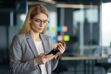 Ofisteki olgun bir iş kadını online işlemler için akıllı telefon ve kredi kartı kullanıyor. Teknoloji, finans ve kolaylığa odaklan. Mesleki çevrede kendine güvenen davranışlar.