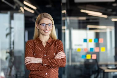 Modern ofiste kollarını kavuşturmuş kendine güvenen olgun bir iş kadını. Puantiyeli tişört giyerek liderliği somutlaştırıyor. Arkasında renkli yapışkan notlar olan cam duvar planlama gösteriyor..