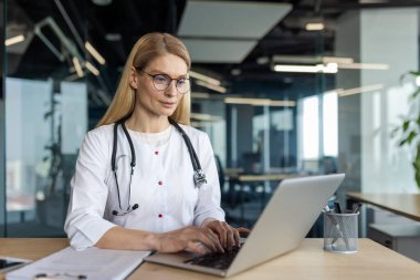 Olgun doktor modern ofiste dizüstü bilgisayara odaklandı. Beyaz önlük, steteskop ve gözlük takıyor. Sahne profesyonelliği, teknolojiyi, sağlık hizmetlerini yakalıyor.