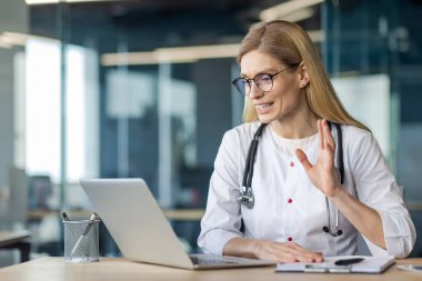 Modern ofisteki dizüstü bilgisayarını kullanarak online danışmada olgun bir kadın doktor. Steteskop takarak sıcak jestler yapıyor. Teletıp, teknoloji, sağlık hizmetleri iletişimi kavramı.