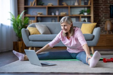 Üst düzey kadın dizüstü bilgisayarla yoga yapıyor, gülümsüyor ve mindere yaslanıyor, ev zindeliğini, sağlığını ve aktif yaşam tarzını vurguluyor. Parlak, rahat oturma odası konforu arttırır..