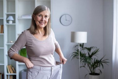 Yaşlı kadın gururla oturma odasında büyük boy pantolon sergiliyor kilo verme başarısını ve mutluluğunu sergiliyor. Kendine güveni kişisel başarısını, sağlığını, sağlıklı yaşam tarzını yansıtıyor..