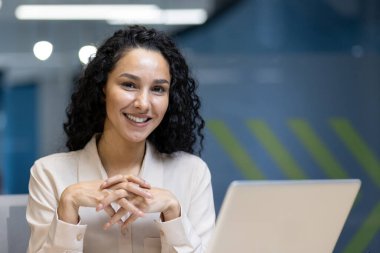 Ofisteki kendine güvenen İspanyol kadın kameraya gülümsüyor. Profesyonel atmosferi kucaklamak, sıcaklığı ve ulaşılabilirliği ifade etmek. Modern işyeri dinamiğinin mükemmel tasviri.