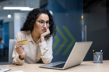 İspanyol kadın kredi kartını ofis ortamında dizüstü bilgisayarla çalışırken tutuyor. Resim, çevrimiçi mali işlemler ile ilişkili stres, belirsizlik ve potansiyel dolandırıcılık risklerini taşır.