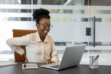 Modern ofis ortamında dizüstü bilgisayarla çalışırken sırtı ağrıyan Afrikalı Amerikalı bir iş kadını. Işıklar teknolojide rahatsızlık yaratıyor. Masanın yanındaki akıllı telefon..