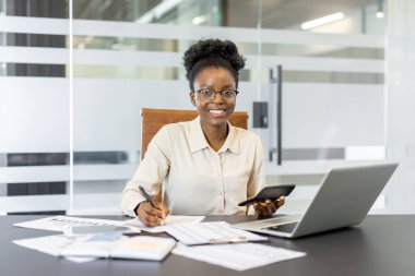 Hesap makinesi, telefon, dizüstü bilgisayar ve finansal belgeleri olan kendinden emin bir Afrikalı Amerikalı iş kadını. Profesyonelliği, finans uzmanlığını, verimliliği gösteriyor. İş ve finans konuları için ideal.