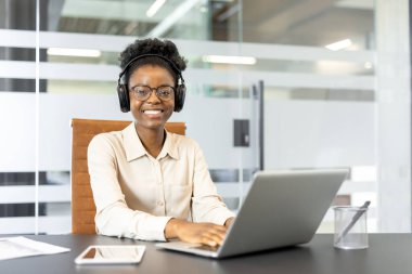 Kulaklık takan Afro-Amerikalı iş kadını modern ofis ortamında dizüstü bilgisayarla çalışıyor. Güvenle gülümsüyor, bağlılık ve keyif ile birden fazla iş yapıyor. Şirket ortamı teşvik eder