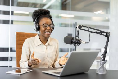 Kulaklıklı Afro-Amerikalı iş kadını podcast seansına katılıyor. Dizüstü bilgisayar ve profesyonel mikrofon kullanarak ofisin görüşlerini paylaşıyor. Ticari iletişim temaları için ideal.