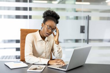 Afro-Amerikalı iş kadını ofis masasında dizüstü bilgisayar ve belgelerle stresli hissediyor. İş yerinde yorgunluk çeken, kafa tokuşturan, yoğun iş günlerinde hayal kırıklığı yaşayan bir kadın..