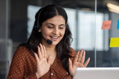 Modern ofis masasındaki sanal toplantıya hevesle katılan, kulaklık takan Latin iş kadını. Gülümseyen ve dinamik, iletişim ve iş alanındaki bağlantıyı ifade eden.