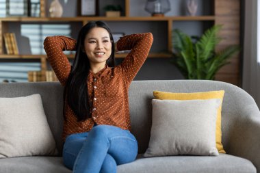 Kanepede oturan gülümseyen kadın, kendini rahat ve mutlu hissediyor. Eller başının arkasında, huzur ve memnuniyet yayıyor. Puantiyeli tişört ve kot pantolon giyiyordu. Modern ev atmosferi.