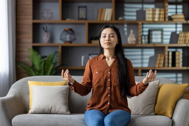 Kanepede meditasyon yapan Asyalı kadın, gözleri kapalı, sükunet ve farkındalığı kucaklayan. Rastgele giyinmiş, rahat bir şekilde oturuyor. Oturma odası düzeni huzurlu ve rahat bir atmosfer sağlar..