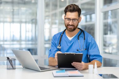 Mavi önlüklü doktor modern ofiste panoya yazarken gülümsüyor. Dizüstü bilgisayarla çalışıyor, profesyonel sağlık ortamını gösteriyor ve tıp alanına kendini adamış..