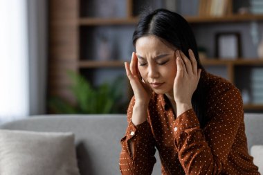 Stres yaşayan Asyalı bir kadın. Kanepede gözleri kapalı, kahverengi benekli tişört giymiş bir şekilde oturuyor. Yüz ifadesi rahatsızlık gösteriyor, baş ağrısı ya da hayal kırıklığı..