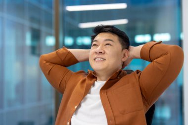 Asyalı işadamı, ellerini başının arkasına koyup rahatça gülümsüyor. Modern arkaplana karşı mesleki tatminin, başarının ve iş hayatının dengesini yakalar.