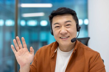 Gülümseyen Asyalı işadamı video görüşmesi için kulaklık kullanıyor. Arkadaş canlısı profesyonel el sallama, iletişim kurma, ofis ortamında modern teknoloji kullanımı..