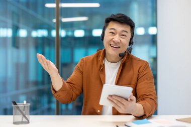 Gülümseyen Asyalı işadamı ofiste sanal toplantı için tablet ve kulaklık kullanıyor. Günlük giysiler, dostça jestler iletişimi ve çalışma alanındaki teknolojiyi vurguladı.