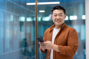 Elinde akıllı telefonla gülümseyen Asyalı işadamı modern ofis ortamında güvenle duruyor. İş hayatında başarı, iletişim ve mesleki güveni temsil eder..