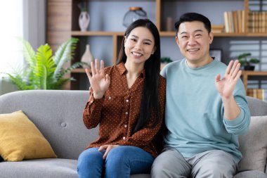 Mutlu Asyalı çift kanepede oturuyor, sıcak bir şekilde gülümsüyor ve el sallıyorlar. Rahat giyimli ev kıyafetleri, sıcaklığı, dostluğu ve birlikteliği ifade eder. Parlak, davetkar oturma odası atmosferi.