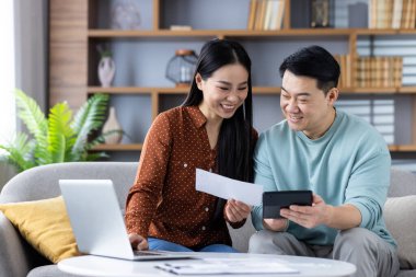 Akıllı telefon ve dizüstü bilgisayar kullanarak gülümseyen Asyalı çift, belgeleri inceliyor. Modern oturma odası düzeni iç çevrede takım çalışması, teknoloji ve mutluluğu yansıtır..