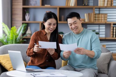 Asyalı çift evde bilgisayarlarıyla belgeleri gözden geçiriyorlar. Resim takım çalışması, mali planlama, aile desteği ve rahat çevre bağlantısı ve işbirliğini yakalar.