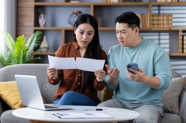 Asyalı çift bilgisayarlı ve akıllı telefonlu finansal belgeleri inceliyor. Erkek ve kadın, ev bütçesi sorunlarını ele alırken kaygılarını dile getiriyorlar. Aile finansmanı, bütçe ve teknoloji entegrasyonu kavramı