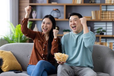 Asyalı çift oturma odasında spor zaferini sevinçle kutluyor. Heyecan ve mutluluk gösteren her ikisi de gülümsüyor. Adamın elinde patlamış mısır kasesi var, iki yumruğu da zafer pozunda, koltukta oturuyor..