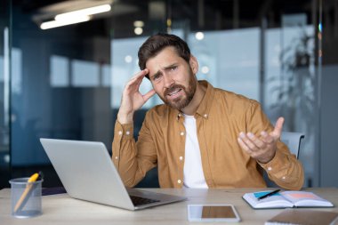 Olgun iş adamı dizüstü bilgisayarı, telefonu ve defteriyle masada oturuyor. Kafa karışıklığı ya da hayal kırıklığını ifade ediyor. Modern ofiste profesyonel ortam. İş durumlarında stresi vurguluyor.