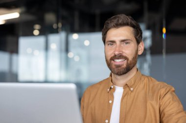 Olgun iş adamı dizüstü bilgisayarla ofiste gülümsüyor. Sıradan tişörtler hem rahat hem de profesyonel davranışlar sergiliyor. Kendine güvenen ifade, olumlu tavır ve iş ortamında başarı öneriyor.
