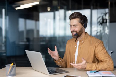 Ofisteki olgun iş adamı bilgisayar ve kulaklık kullanarak video görüşmesine katılıyor. Bağlı ve konuşkan görünüyor, sanal toplantı sırasında el kol hareketi yapıyor, uzaktan çalışma dinamiklerini vurguluyor..