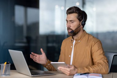Video konferans için bilgisayar ve tablet kullanan, kulaklık takan olgun bir işadamı. İletişimle meşgul, birden fazla görev becerisi gösteriyor. Profesyonel ayarlar işi güçlendirir
