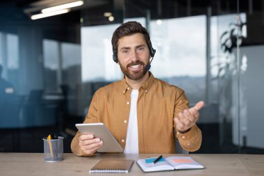 Video görüşmesi sırasında, tablet ve kulaklık kullanarak ofiste oturan profesyonel bir iş adamı. Jest, misafirperver havayı uzatır. Modern iletişim, teknoloji, işyeri etkileşimini kavramsallaştırır.