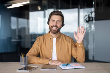 Ofisteki gülümseyen adam, kulaklık takıyor, video çağrısı yapıyor, el sallıyor. Dostça iletişim sağlandı. Defter, tablet ve kalemle birlikte çalışma masasında, profesyonel çalışma ortamının iletilmesi.