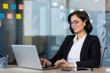 Modern ofiste dizüstü bilgisayarla çalışan bir iş kadını. Gözlük takan profesyonel bir kadın ve resmi kıyafetler görevlere odaklandı. Masadaki eşyalar arasında telefon ve not defteri, üretken ayarların geliştirilmesi yer alıyor.