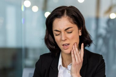 İş kadını iş yerinde diş ağrısı çekiyor, acı ve rahatsızlığını yüz ifadesi ile ifade ediyor. Profesyonel kadın, ofis kıyafetiyle çenesine dokunuyor, diş ağrısı çekiyor..