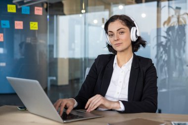 Takım elbiseli, bilgisayar kullanan ve modern ofiste kulaklık takan kendine güvenen bir iş kadını. Profesyonel patron çevrimiçi iletişimde, işine odaklanmış. Kulaklıklar çoklu görev yeteneğini gösteriyor