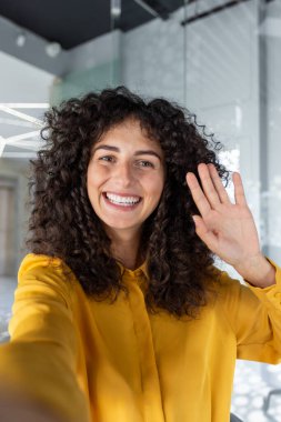 Latin Amerikalı iş kadını sarı tişört giyiyor. Ofiste sanal toplantı sırasında gülümsüyor ve el sallıyor. Kıvırcık saçlı kadın dostluk ve pozitiflik yayıyor. Akıllı telefonu bağlantı için kullanıyor..