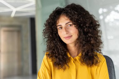 Sarı bluz giyen kıvırcık saçlı Latin Amerikalı iş kadını. Modern ofiste profesyonelliği, kendine güveni ve ulaşılabilirliği ifade eder. Şirket, liderlik, iş konuları için ideal.