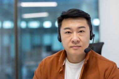 Ofisteki Asyalı adam kulaklık takıyor, profesyonelliğini, iletişim becerilerini ve iş ortamını gösteriyor. Resim, iş etkileşimi sırasında sakin ve kendinden emin tavırları yakalar.