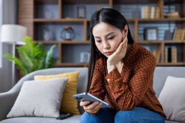 Bluz giymiş Asyalı bir kadın kanepeye oturmuş elinde akıllı bir telefonla düşünceli ve düşünceli görünüyor. Ahşap raflı ev dekoru modern ve rahat atmosfere ekleniyor. Anahtar kelimeler: kadın, telefon, düşünceli.