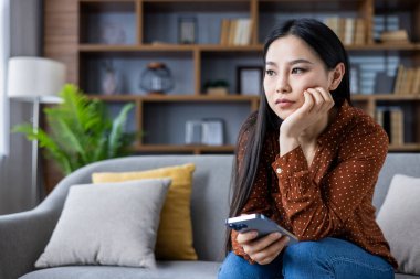 Genç Asyalı kadın koltukta oturuyor, telefonu tutuyor, düşünceli görünüyor. Günlük kıyafet rahat bir ortam sağlıyor. İfade, tefekküre ya da üzüntüye işaret eder, belki de evdeki kişisel yansımaya..