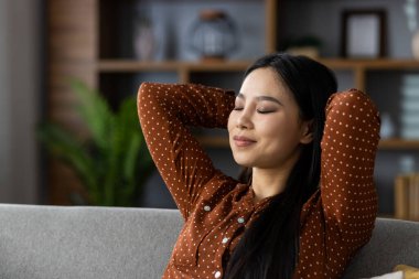 Asyalı kadın kanepede gözleri kapalı, elleri başının arkasında dinleniyor. Huzurlu ifade evde sükunet ve sükunet gösterir. Sıradan polka noktalı tişört sükunet anına stil katar.