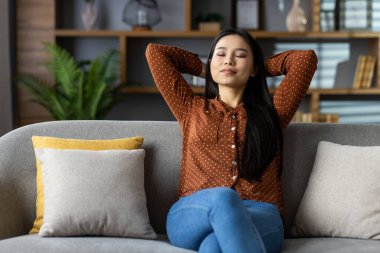 Asyalı bir kadın evinde kanepede dinleniyor, gözleri kapalı, elleri başının arkasında. Huzur ve huzuru kucaklıyor, rahat oturma odasında sessiz anların tadını çıkarıyor. Rahatlama, sükunet kavramı.