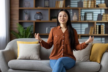 Rahat bir Asyalı kadın evdeki kanepede meditasyon yapıyor, huzurlu ve huzurlu bir ifadeyle. Farkındalık ve kendine bakmakla meşgul, sükunet ve gevşemeyi somutlaştıran.