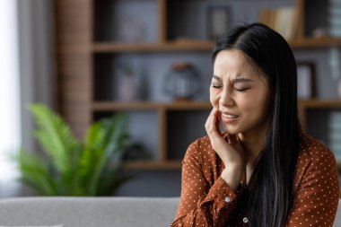 Asyalı kadın evde diş ağrısı belirtileri göstererek yanağını rahatsızlık içinde tutuyor. Kahverengi puantiyeli tişört, koltukta oturmak, arka plan ifadeye odaklanmak için bulanık.