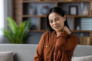 Kahverengi benekli tişörtlü Asyalı kadın evde boynu ağrıyor. Kanepede gözleri kapalı ve elleri boynunda oturuyor. Rahatsızlığı yansıtıyor. Oturma odası arka planda görünüyor.