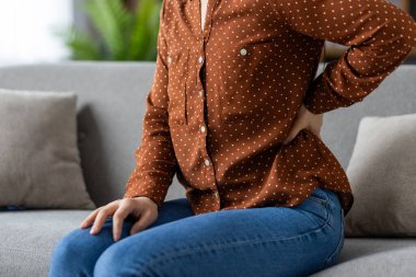 Sırtı ağrıyan bir kadın kanepede oturuyor, belini tutuyor. Kahverengi benekli gömlek ve kot pantolon giyiyor. Rahatsızlık gösteriyor. Ağrı ya da sakatlık gibi sağlık sorunlarına işaret ediyor..