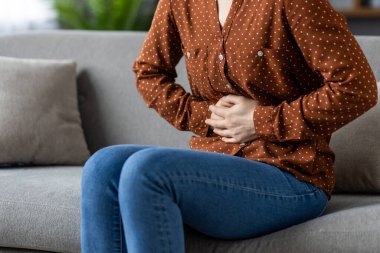 Karnı ağrıyan genç bir kadın koltukta oturuyor, karnını tutuyor, rahatsızlık gösteriyor. Kahverengi benekli gömlek, mavi kot pantolon giyer. Sağlık, acı ve rahatsızlık kavramları taşır..